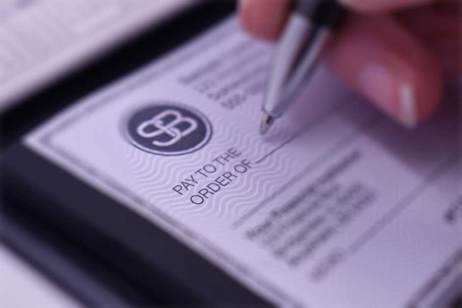 A close-up of a hand holding a pen over a modern, professionally designed check, focusing on the &ldquo;Pay to the Order Of&rdquo; section with subtle security patterns and a clean, polished layout.